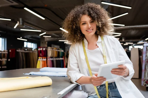 Zufriedene Verkäuferin hält digitales Tablet in der Nähe von Stoffrollen auf Schreibtisch im Textilgeschäft — Stockfoto