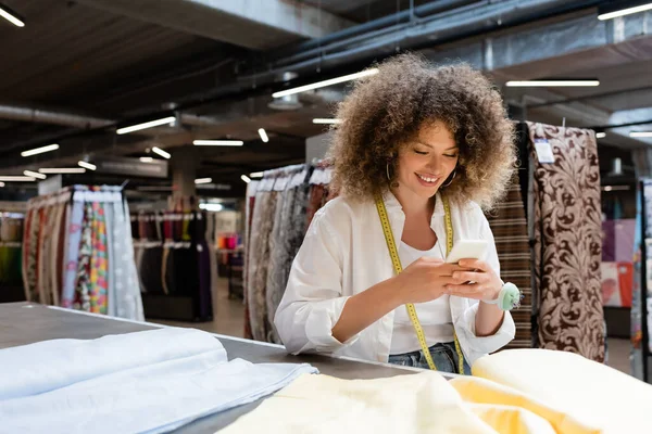 Vendeuse positive utilisant un smartphone près du tissu sur le bureau dans un magasin de textiles — Photo de stock