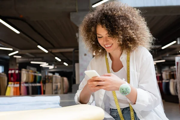 Zufriedene Verkäuferin mit Smartphone in der Nähe von Stoffrollen auf dem Schreibtisch im Textilgeschäft — Stockfoto