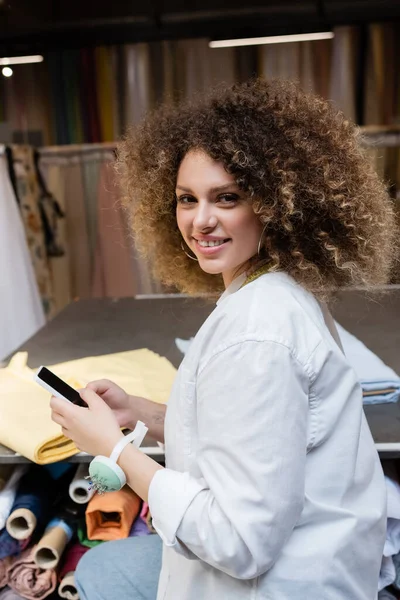 Vendedora positiva usando teléfono inteligente con pantalla en blanco en la tienda textil - foto de stock