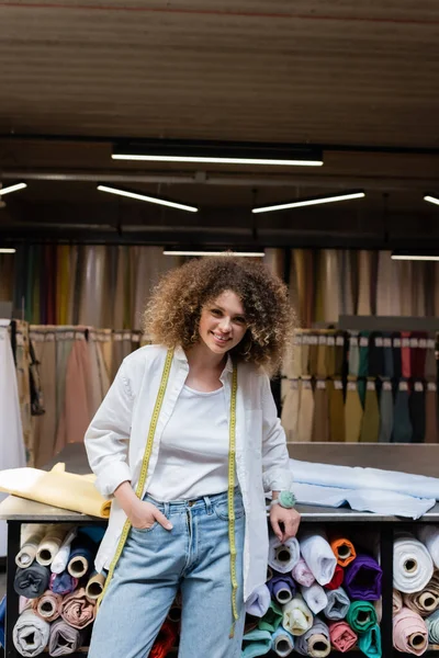 Положительная продавщица с вьющимися волосами, стоящая с рукой в кармане возле стойки с рулонами ткани — стоковое фото