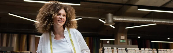 Cheerful saleswoman with curly hair standing in textile shop, banner — Stock Photo