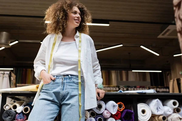 Low angle view of smiling saleswoman with curly hair standing with hand in pocket near rack with fabric rolls — Stock Photo