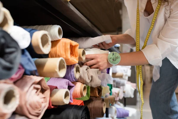 Vista recortada de la vendedora elegir entre rollos de tela de colores en los estantes de la tienda textil - foto de stock