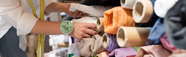 Vue recadrée de vendeuse au choix entre des rouleaux de tissu colorés sur les étagères de la boutique de textiles, bannière — Photo de stock