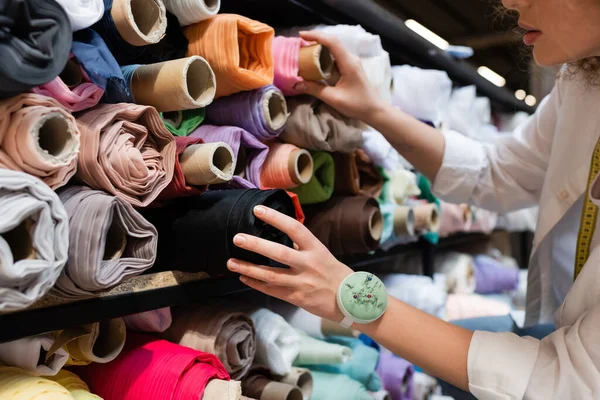 Vista recortada del vendedor elegir entre rollos de tela de colores en los estantes de la tienda textil - foto de stock