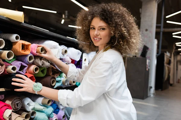 Fröhliche Verkäuferin holt rosa Stoffrolle aus Regal in Textilgeschäft — Stockfoto