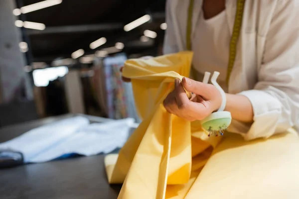 Cropped view of salesperson with needle cushion on hand holding yellow fabric while working in textile shop — Stock Photo