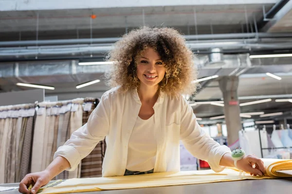 Vendedora alegre com almofada de agulha na mão medindo tecido amarelo na loja têxtil — Fotografia de Stock