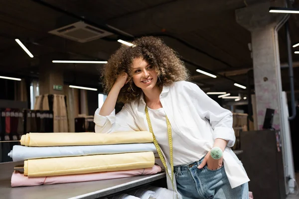 Cheerful saleswoman with needle cushion posing with hand in pocket while leaning on desk with colorful fabric rolls — Stock Photo