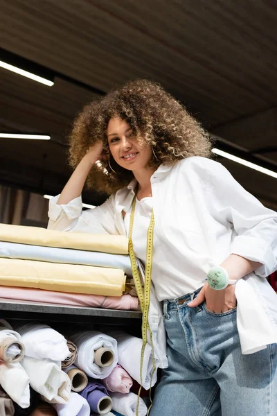 Улыбающаяся продавщица с игольчатой подушкой, позирующая с рукой в кармане, опираясь на стойку с разноцветными рулонами ткани — стоковое фото