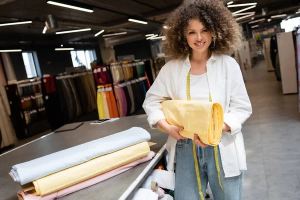 Positive saleswoman holding pastel yellow fabric roll in textile shop — Stock Photo