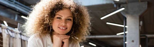 Pleased saleswoman with curly hair looking at camera in textile shop, banner — Stock Photo