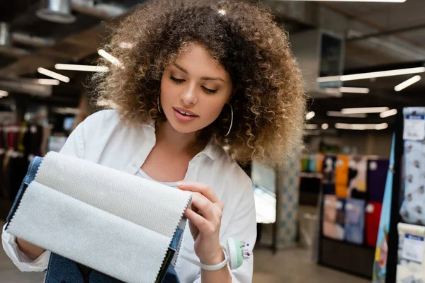 Mulher encaracolado com almofada de agulha na mão escolher entre diferentes amostras de tecido na loja têxtil — Fotografia de Stock