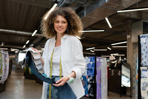 Fröhliche Verkäuferin mit Nadelkissen in der Hand, die bunte Stoffmuster im Textilladen hält — Stockfoto