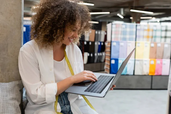 Щасливий продавець з вимірювальною стрічкою за допомогою ноутбука під час роботи в текстильному магазині — стокове фото