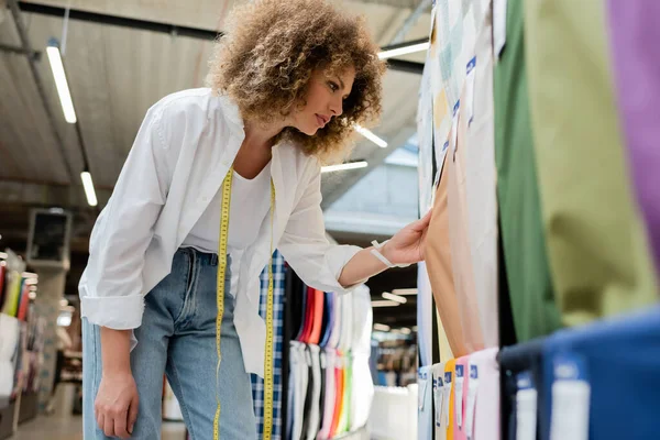 Side view of curly saleswoman with measuring tape choosing among patterned fabric in textile store — Stock Photo