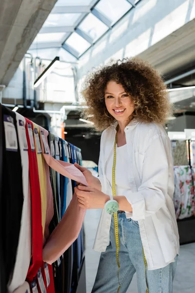 Curly saleswoman smiling at camera while choosing from various fabric in textile shop — Stock Photo