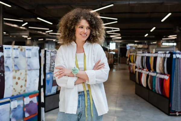 Curly and cheerful saleswoman standing with crossed arms and looking away in textile shop — Stock Photo