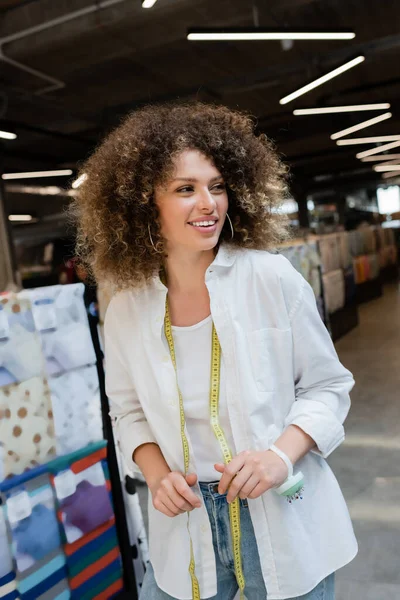 Cheerful and curly saleswoman with measuring tape looking away in textile shop — Stock Photo