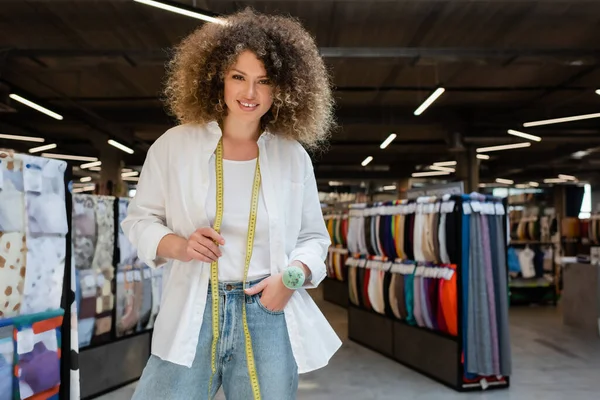 Curly saleswoman with measuring tape and needle cushion standing with hand in pocket of jeans in textile shop — Stock Photo