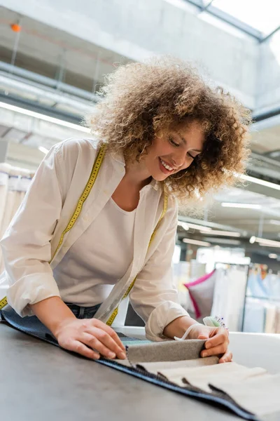 Curly saleswoman with measuring tape looking at fabric samples in textile shop — Stock Photo