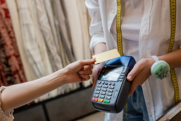 Обрезанный вид продавщицы, держащей считыватель кредитных карт рядом с клиентом в текстильном магазине — стоковое фото