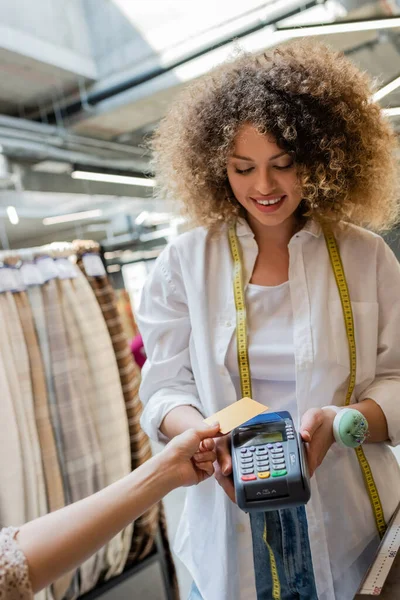 Happy and curly seller with measuring tape holding payment terminal near customer with credit card in textile shop — Stock Photo