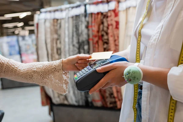 Partial view of saleswoman with needle cushion on hand holding credit card reader near buyer in textile shop — Stock Photo