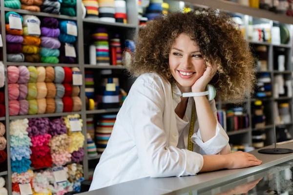 Happy saleswoman with needle cushion on hand looking away near counter and rack with needlework accessories — Stock Photo