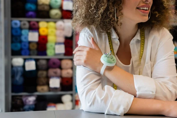 Partial view of smiling saleswoman with measuring tape and needle cushion near counter and blurred rack with yarn skeins — Stock Photo