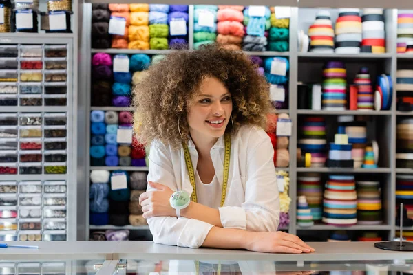 Curly saleswoman with measuring tape and needle cushion looking away near counter and rack with needlework accessories — Stock Photo