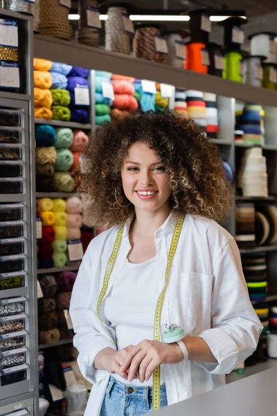 Curly saleswoman with measuring tape smiling at camera near rack with needlework and knitting accessories — Stock Photo