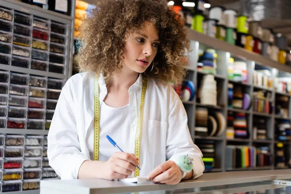 Pretty and curly saleswoman making order near blurred rack with needlework accessories — Stock Photo