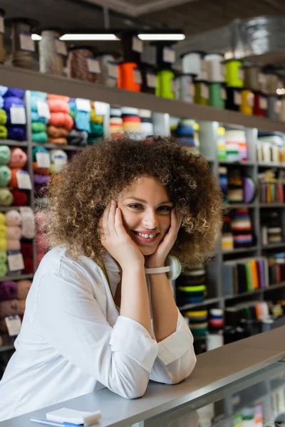Happy and curly saleswoman holding hands near face while looking at camera in textile shop — Stock Photo