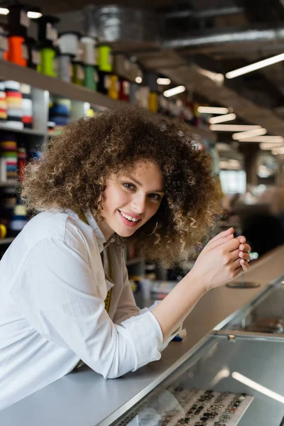 Cheerful saleswoman smiling at camera near counter in textile shop — Stock Photo