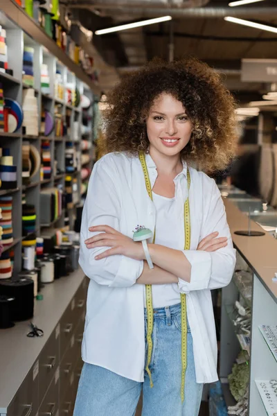 Curly saleswoman with measuring tape and needle cushion standing with crossed arms at counter in textile shop — Stock Photo