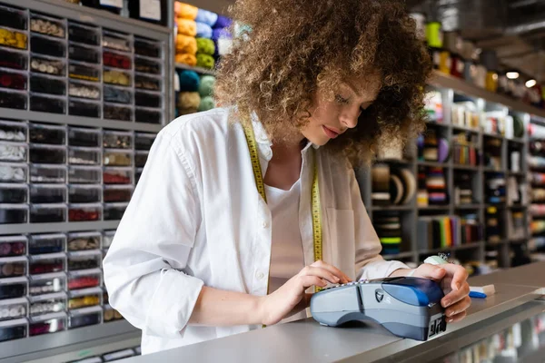 Vendedora joven y rizado que trabaja con lector de tarjetas de crédito en el mostrador de la tienda textil - foto de stock