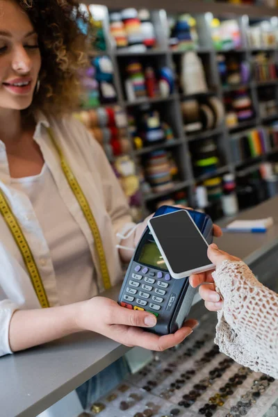 Молодая продавщица с измерительной лентой, держащая платежный терминал рядом с покупателем с мобильным телефоном в текстильном магазине — стоковое фото
