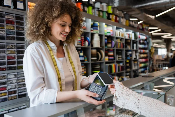 Куртуазна продавниця тримає платіжний термінал поруч з клієнтом з мобільним телефоном в текстильній крамниці — стокове фото