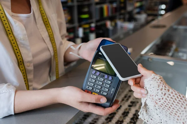 Partial view of woman holding smartphone near payment terminal in hands of seller in textile shop — Stock Photo
