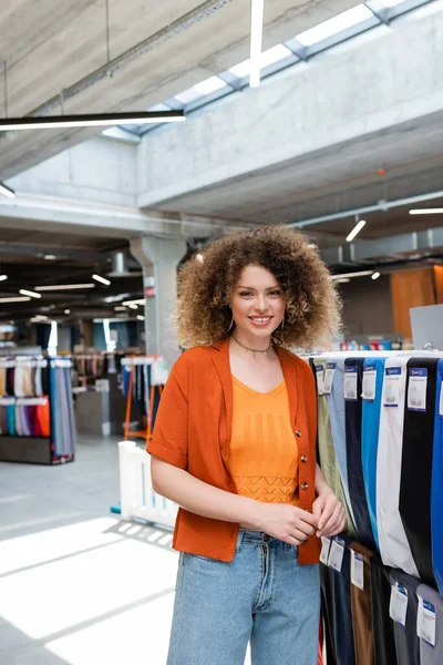 Joyful woman looking at camera near variety of multicolored fabric in textile shop — Stock Photo
