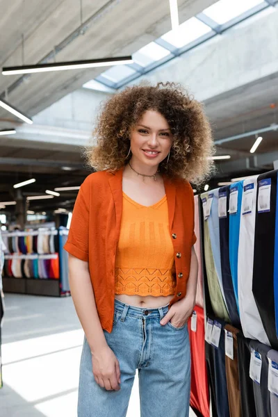 Curly woman standing with hand in pocket and smiling at camera in textile shop — Stock Photo
