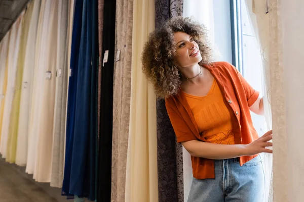 Fröhlich lockige Frau blickt auf verschiedene Gardinen in Textilgeschäft — Stockfoto