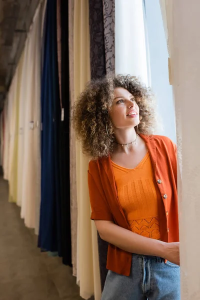 Счастливая молодая женщина с вьющимися волосами выбирает новые занавески в текстильном магазине — стоковое фото