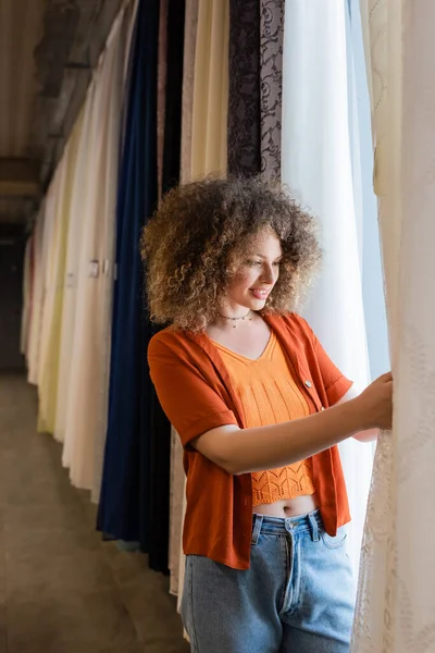 Pretty young woman making choice from variety of curtains in textile shop — Stock Photo