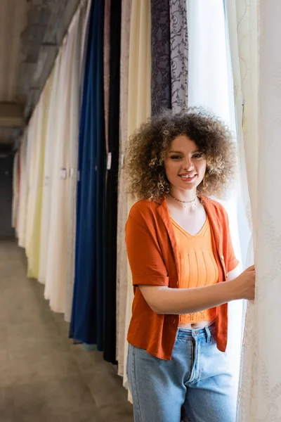 Pretty and curly woman looking at camera near various curtains in textile shop — Stock Photo