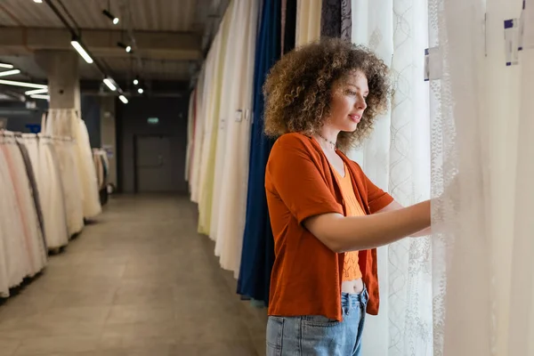 Young and curly woman choosing curtains from assortment in textile shop — Stock Photo