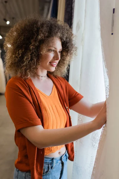 Happy and curly woman looking at white curtains in textile shop — Stock Photo