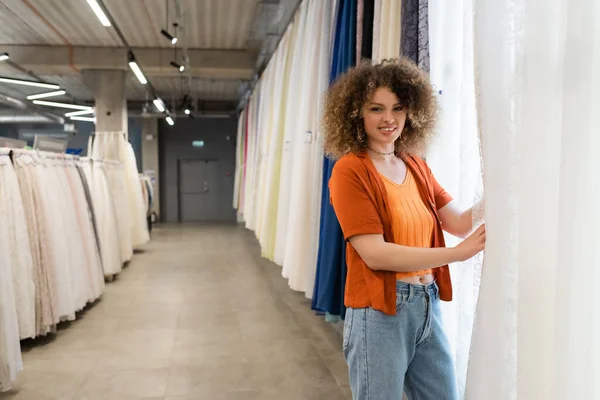 Mulher feliz e encaracolado olhando para a câmera perto variedade de cortinas na loja têxtil — Fotografia de Stock